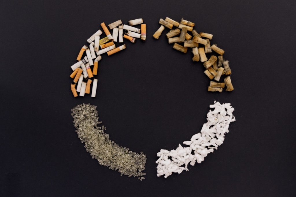 Kreisförmig angeordnete Materialien auf dunklem Hintergrund – Zigarettenreste, Fasern und Kunststoffgranulat als Symbol für Recycling und Upcycling.