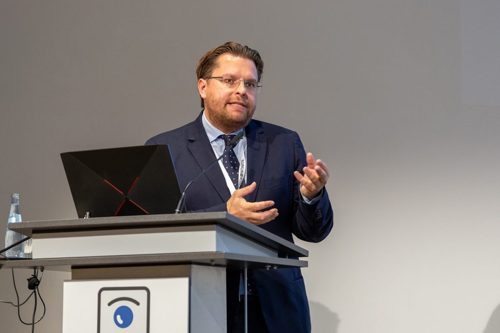 Ein Redner im Anzug spricht am Podium und gestikuliert während einer Präsentation.