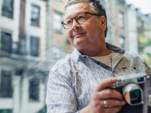 Mann mit Brille und Fotokamera steht im Raum an ein Fenster gelehnt