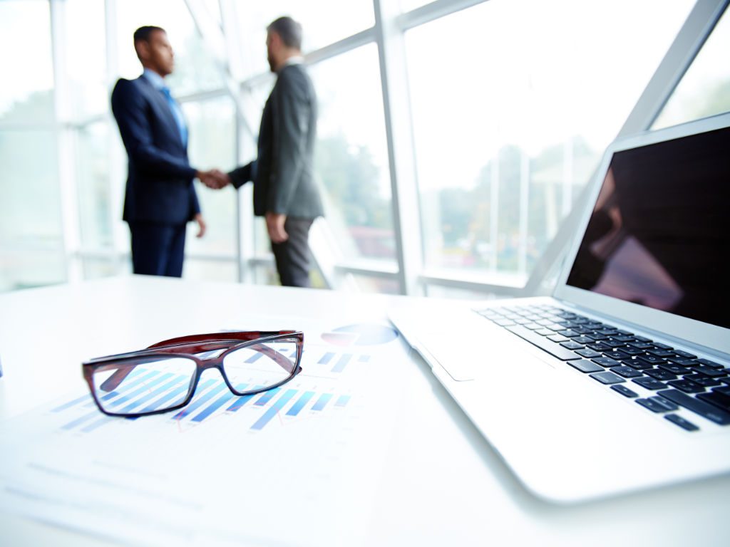 Geschäftlicher Handschlag im Hintergrund, im Vordergrund liegt eine Brille auf dem Tisch