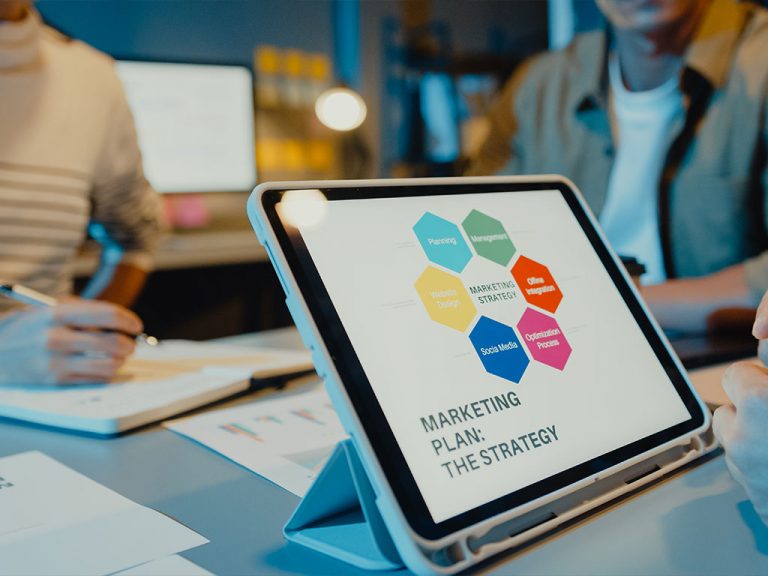 Tablet auf einem Besprechungstisch zeigt eine Grafik zu einer Marketingstrategie. Im Hintergrund arbeiten Personen an Unterlagen und Bildschirmen in einer Büroumgebung zusammen.